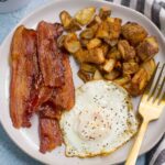 How to Cook Crispy Bacon - showing bacon on white plate with potatoes and a fried egg.