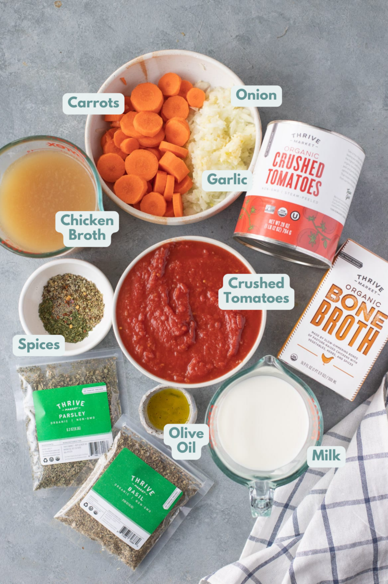 Ingredients for tomato basil soup in white bowls: olive oil, onion, garlic, carrots, canned crushed tomatoes, broth, dried basil, dried oregano, dried parsley, pepper, milk, fresh basil (optional)