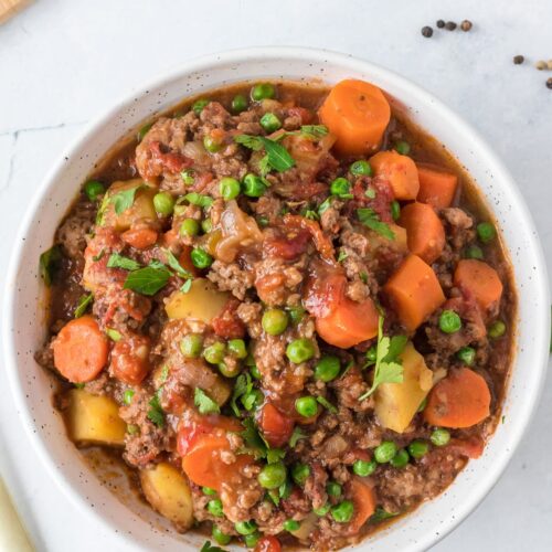 A close up of a bowl of ground beef stew on a white background with a spoon and ingredients off to the side