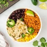 an overhead shot of salsa verde chicken in a white bowl with black beans, corn, and rice with cilantro, jalapeños and limes on the side