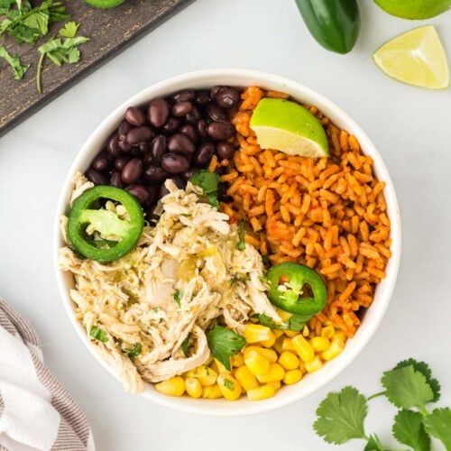 an overhead shot of salsa verde chicken in a white bowl with black beans, corn, and rice with cilantro, jalapeños and limes on the side