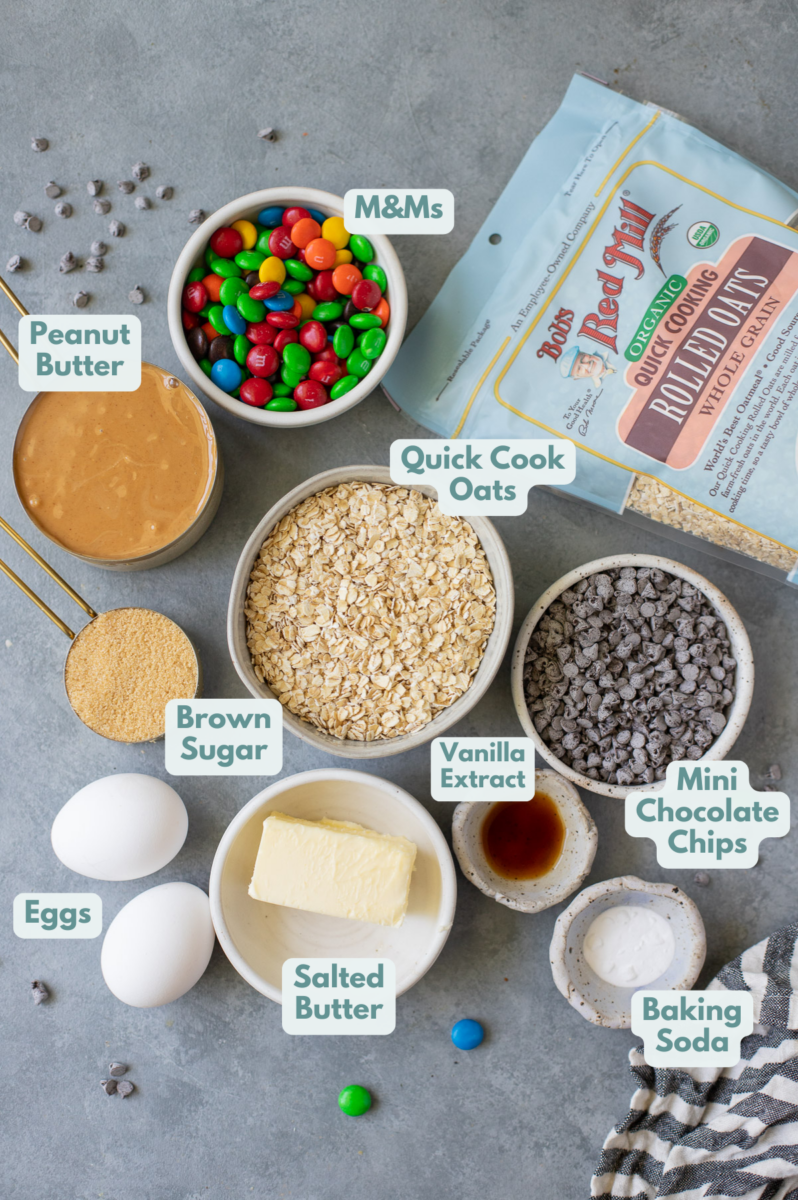 Ingredients for healthy monster cookies in white bowls: salted butter, brown sugar, creamy peanut butter, eggs, vanilla extract, baking soda, Bob's Red Mill Organic Quick Cook Oats, mini chocolate chips, m&ms