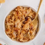 An overhead shot of a white bowl filled with caramelized onions with a gold fork in it on a marble background