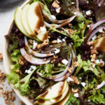 An overhead shot of an apple walnut salad with vinaigrette on top in a white bowl on a white background