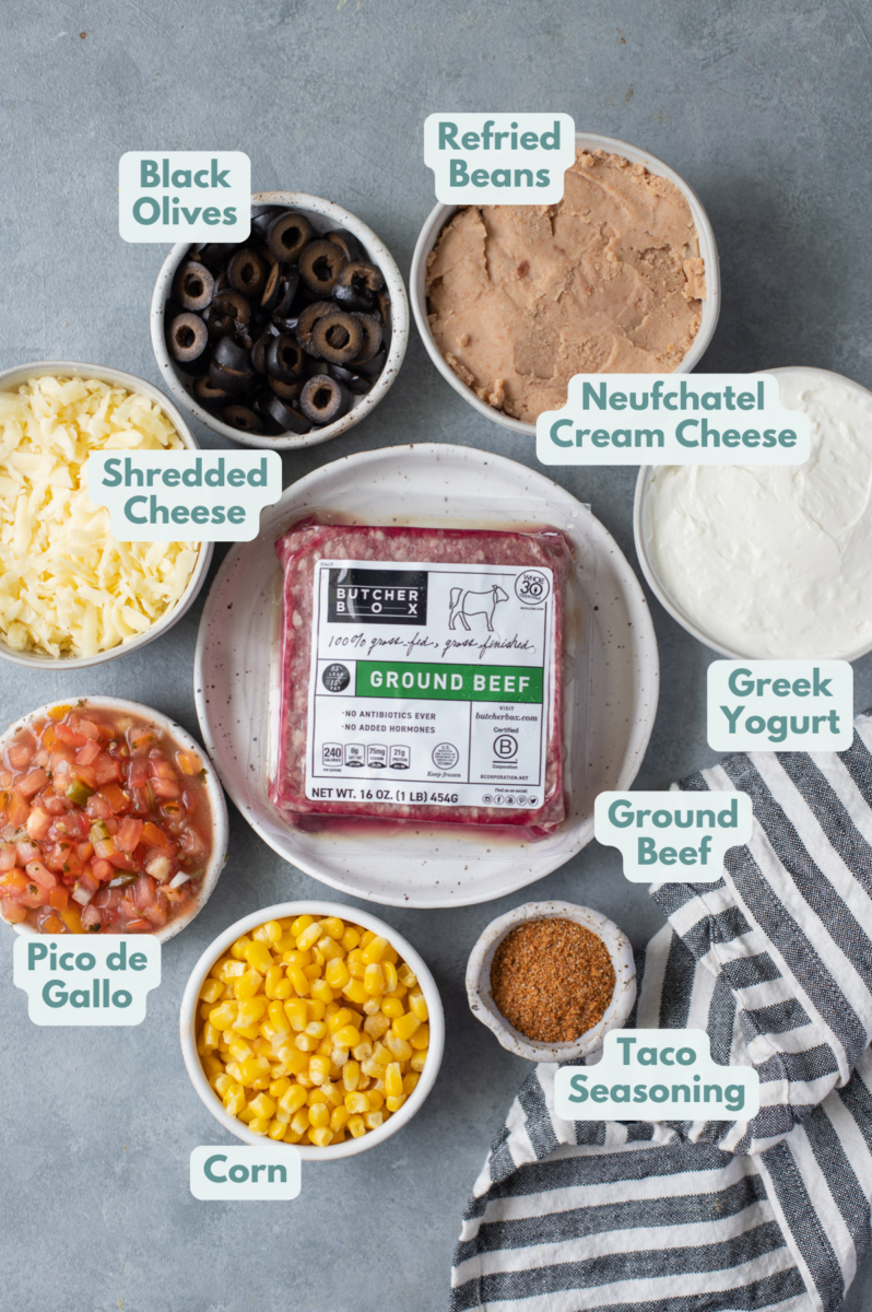 Ingredients for baked 7 layer dip in white bowls: ground beef, taco seasoning, greek yogurt, neufchatel cream cheese, refined beans, corn, black olives, shredded cheddar cheese, pico de gallo
