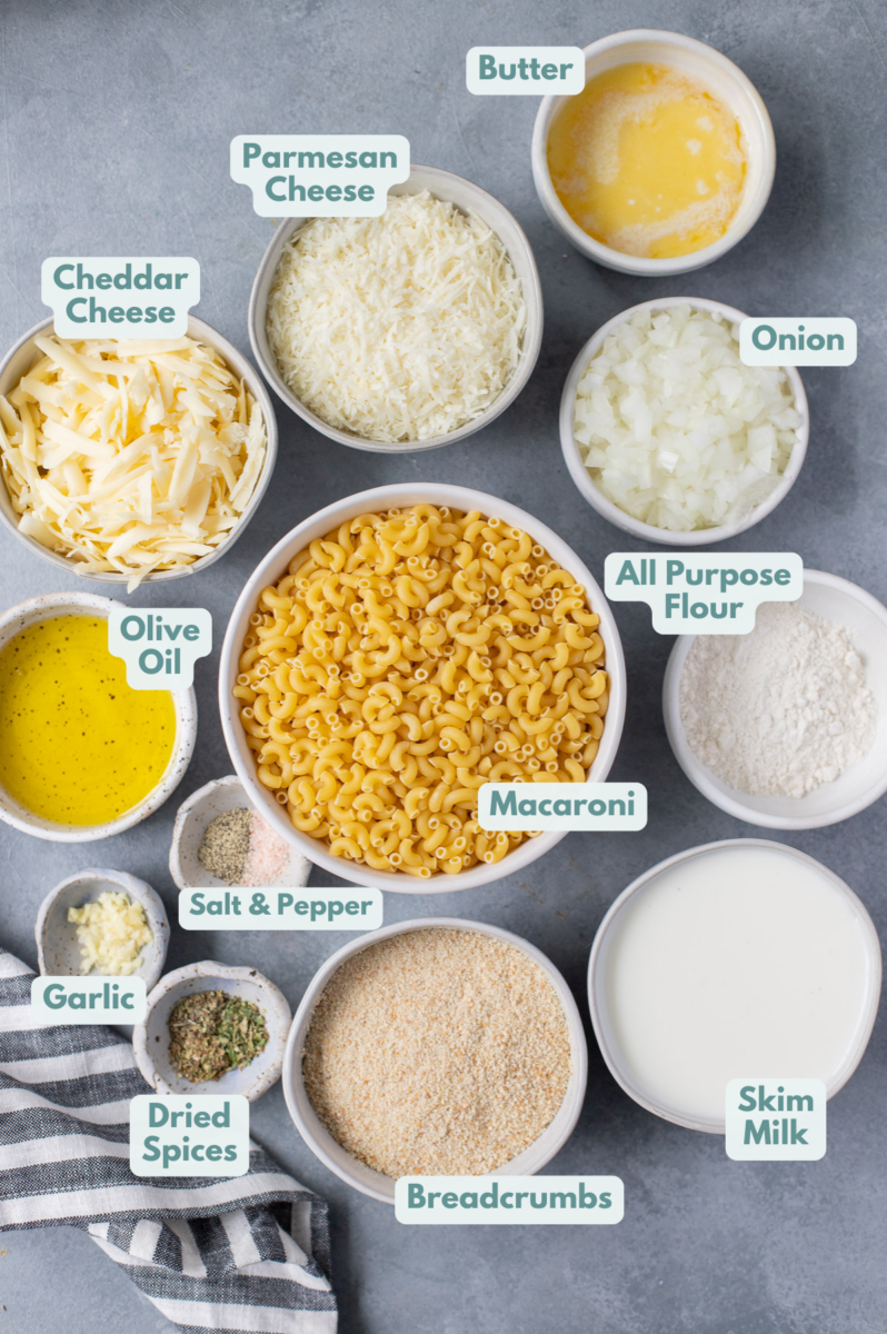 Ingredients for healthy mac and cheese in white bowls: elbow macaroni pasta, olive oil, onion, all purpose flour or gluten free, skim milk, garlic, shredded cheddar cheese, parmesan cheese, salt & pepper, breadcrumbs, dried basil, dried parsley, dried oregano, melted butter