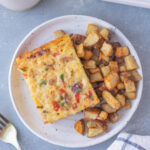 An overhead shot of a white plate with a piece of bacon breakfast casserole and roasted potatoes with another casserole dish, a mug of coffee, and fork on the side