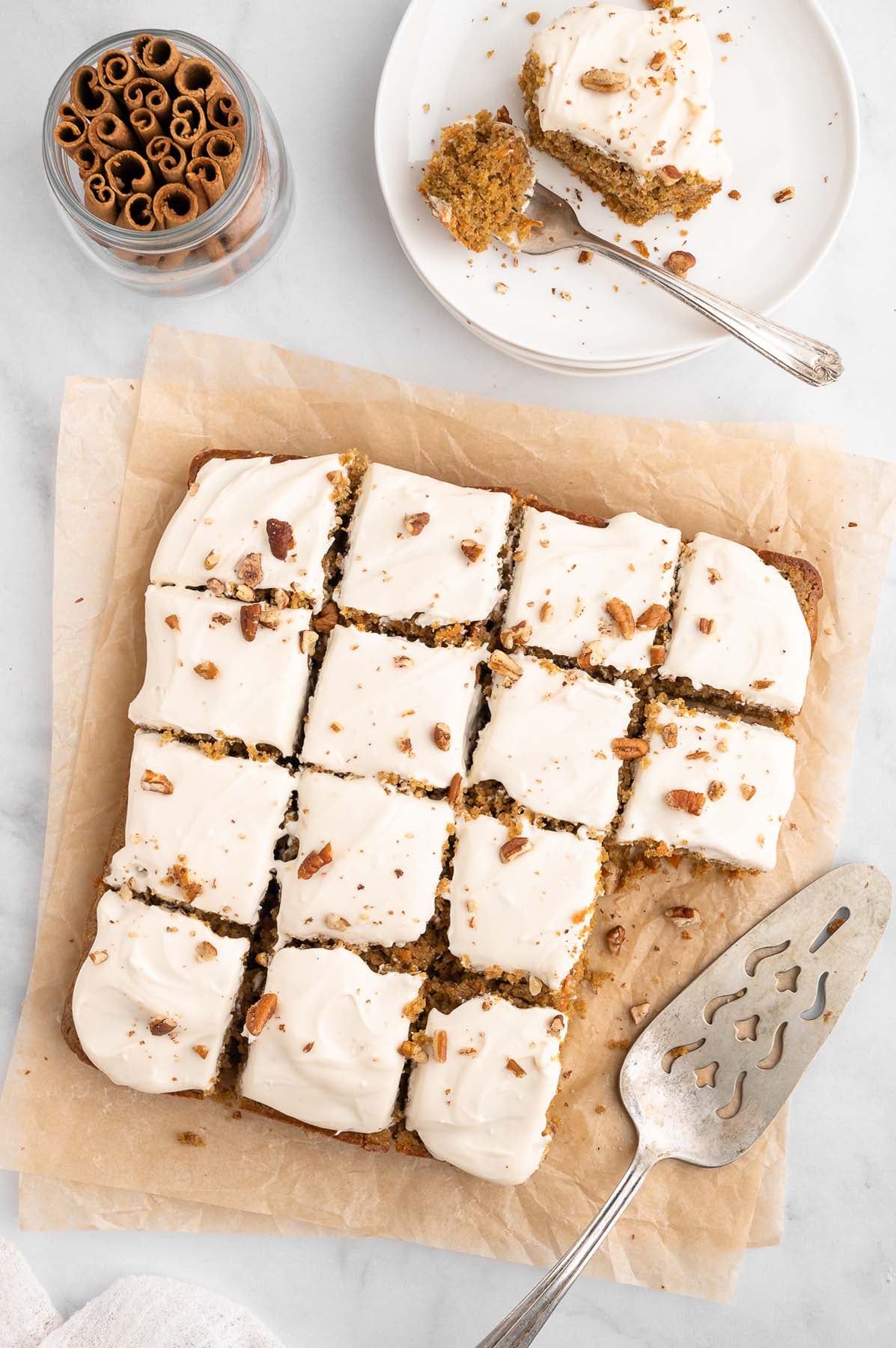 An overhead shot of healthy carrot cake sliced into sixteen squares, with two pieces missing. A serving spatula sits next to the cake.