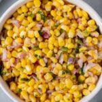 An overhead shot of a white bowl with corn salsa with a sliced lime and cilantro on the side on a grey background