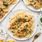 An overhead shot of chicken piccata on a white plate with a fork, lemons, another plate and platter on the side