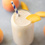 A peach smoothie in a clear glass with a blue and white striped straw in it garnished with a peach slice and more sliced peaches in the background
