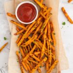 An overhead shot of baked sweet potato fries on a parchment lined platter with a white bowl of ketchup on the platter on a grey background