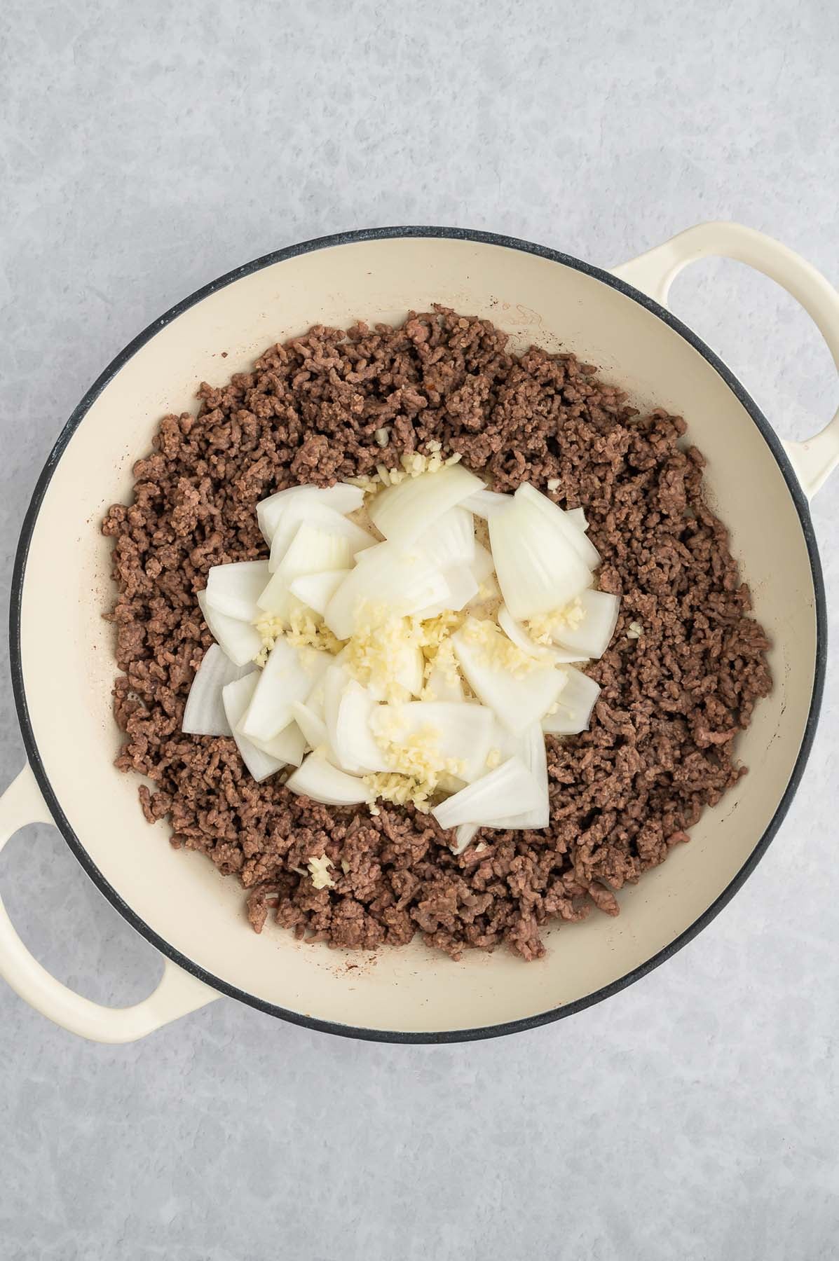 Overhead view of cooked ground hamburger pushed to the edges of a dutch oven with sliced onions and garlic in the middle.