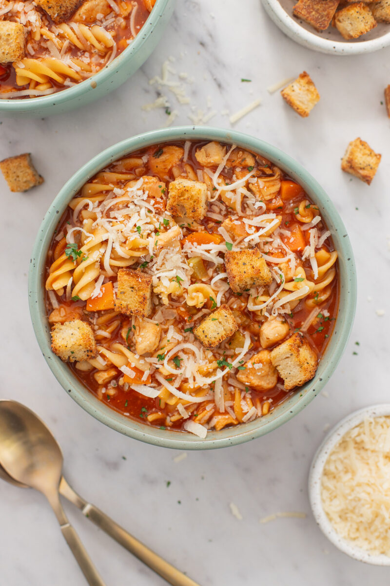 A bowl of chicken parmesan soup topped with croutons and more croutons and spoons spread on a counter.