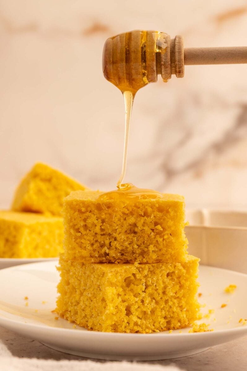 Two pieces of healthy honey cornbread stacked on each other on a plate with a honey dipper drizzling honey on top. 