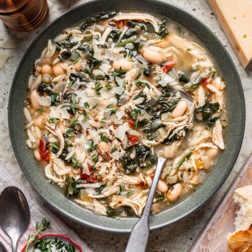 Tuscan Chicken Soup in a grey bowl with a spoon in it and parmesan cheese rind on the side.