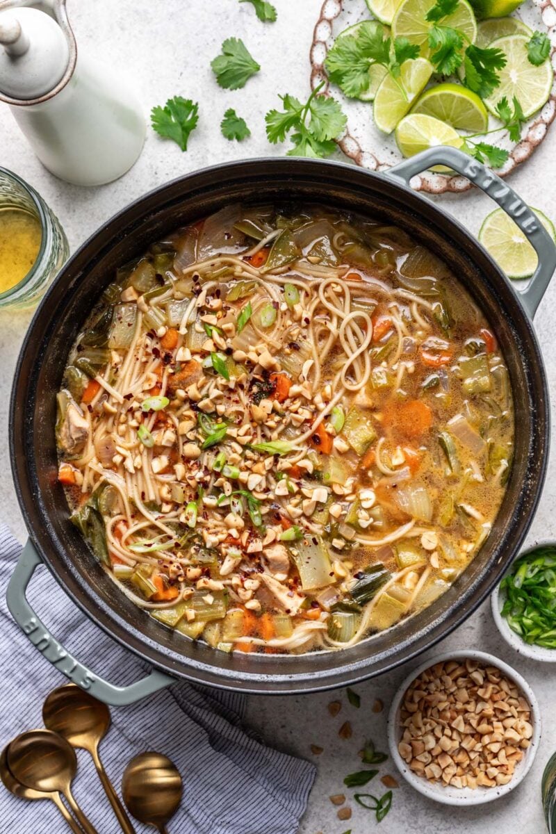 A large dutch oven pot with chicken rice noodle soup and limes and chopped peanuts on the side.