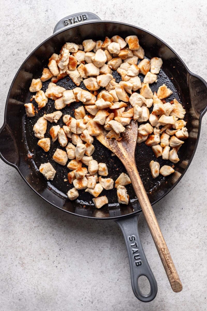 Chicken pieces browned in a cast iron skillet with a wooden spoon in it. 