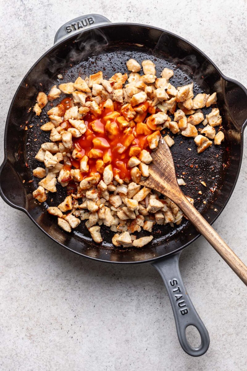Browned chicken in a cast iron skillet with buffalo sauce poured in before being mixed together with a wooden spoon in it.