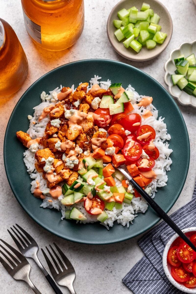 A bowl of rice topped with buffalo chicken, veggies and topped with buffalo sauce and bowls of veggies on the side.