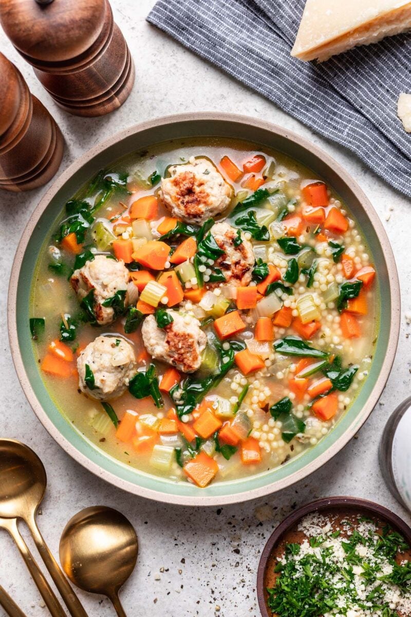 Chicken Meatball Soup in a large bowl with a bowl of herbs and cheese and more spoons on the side.