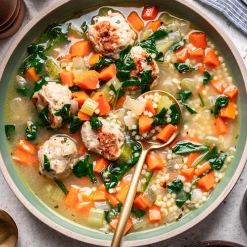 Chicken Meatball Soup in a bowl with a gold spoon in it with a bowl of herbs and cheese on the side.