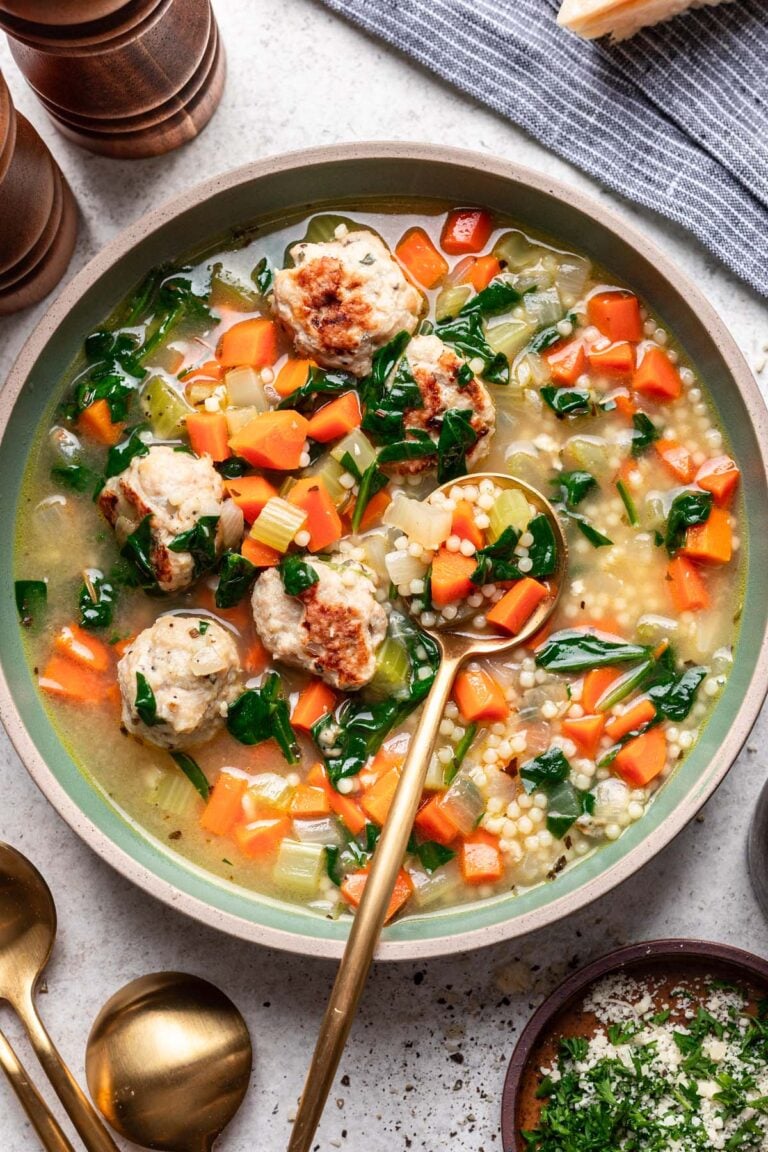 Chicken Meatball Soup in a bowl with a gold spoon in it with a bowl of herbs and cheese on the side.
