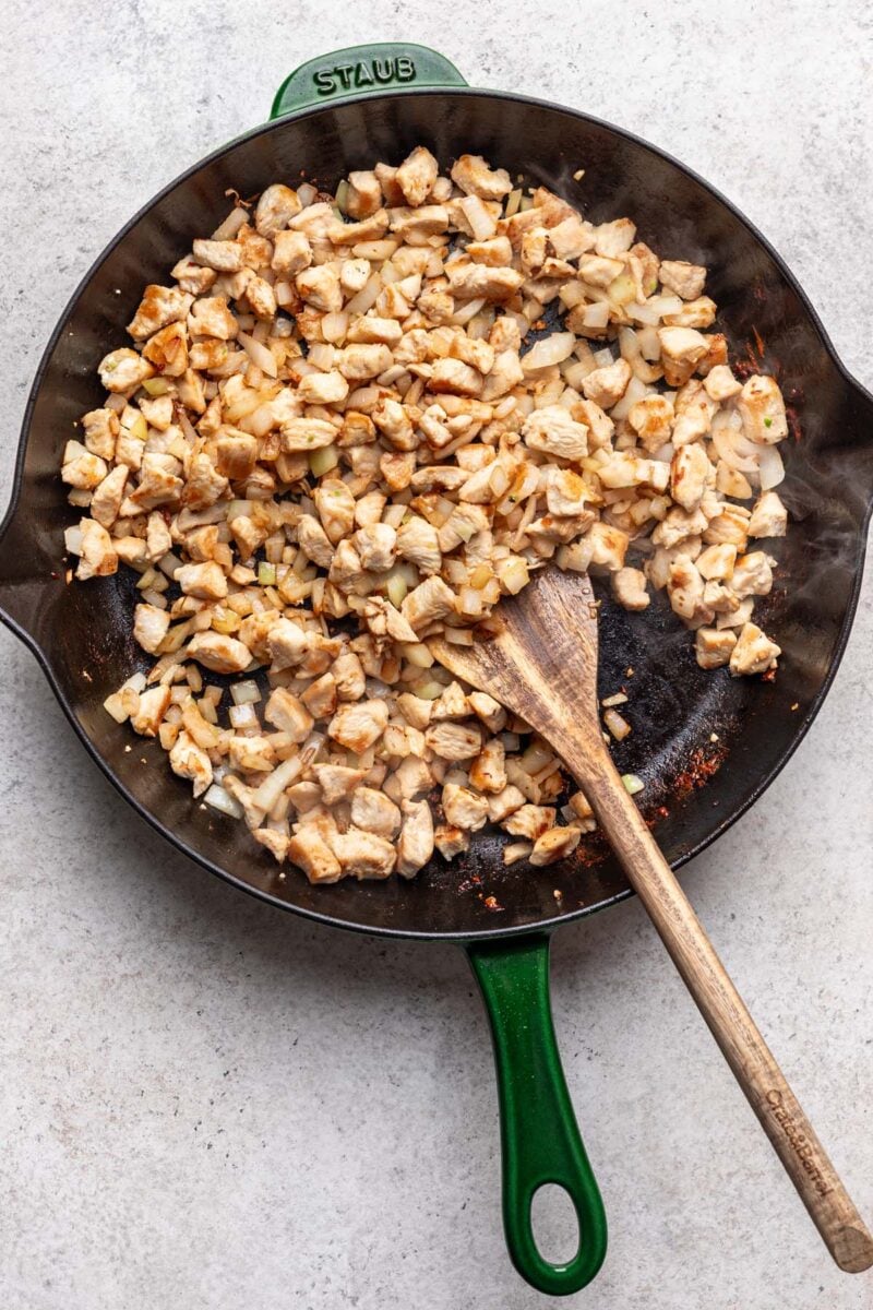 Cubed chicken browned in a cast iron skillet with onions and garlic and a wooden spoon mixing them. 