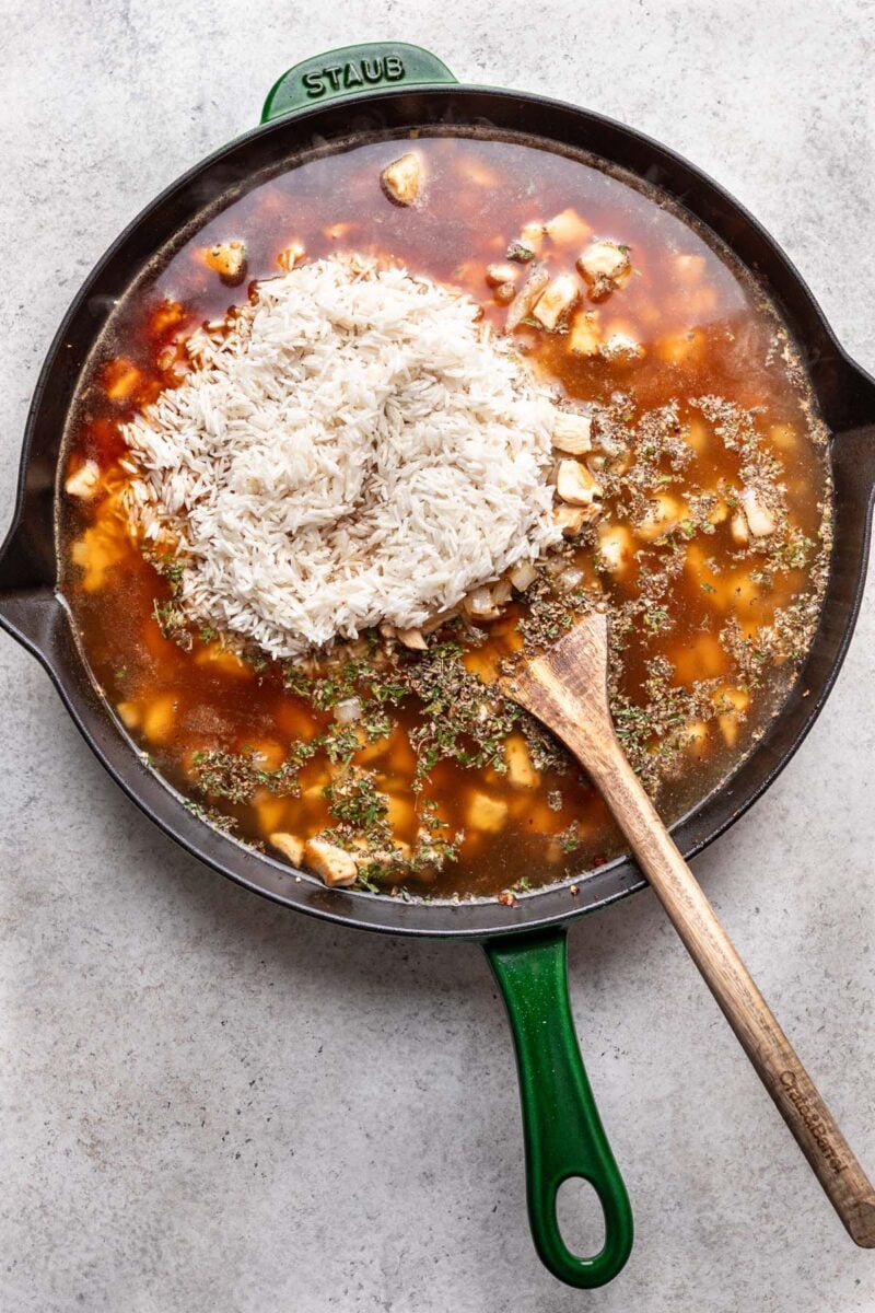 Chicken broth and dried rice in a cast iron skillet with cooked chicken before being mixed together with a wooden spoon resting in it. 