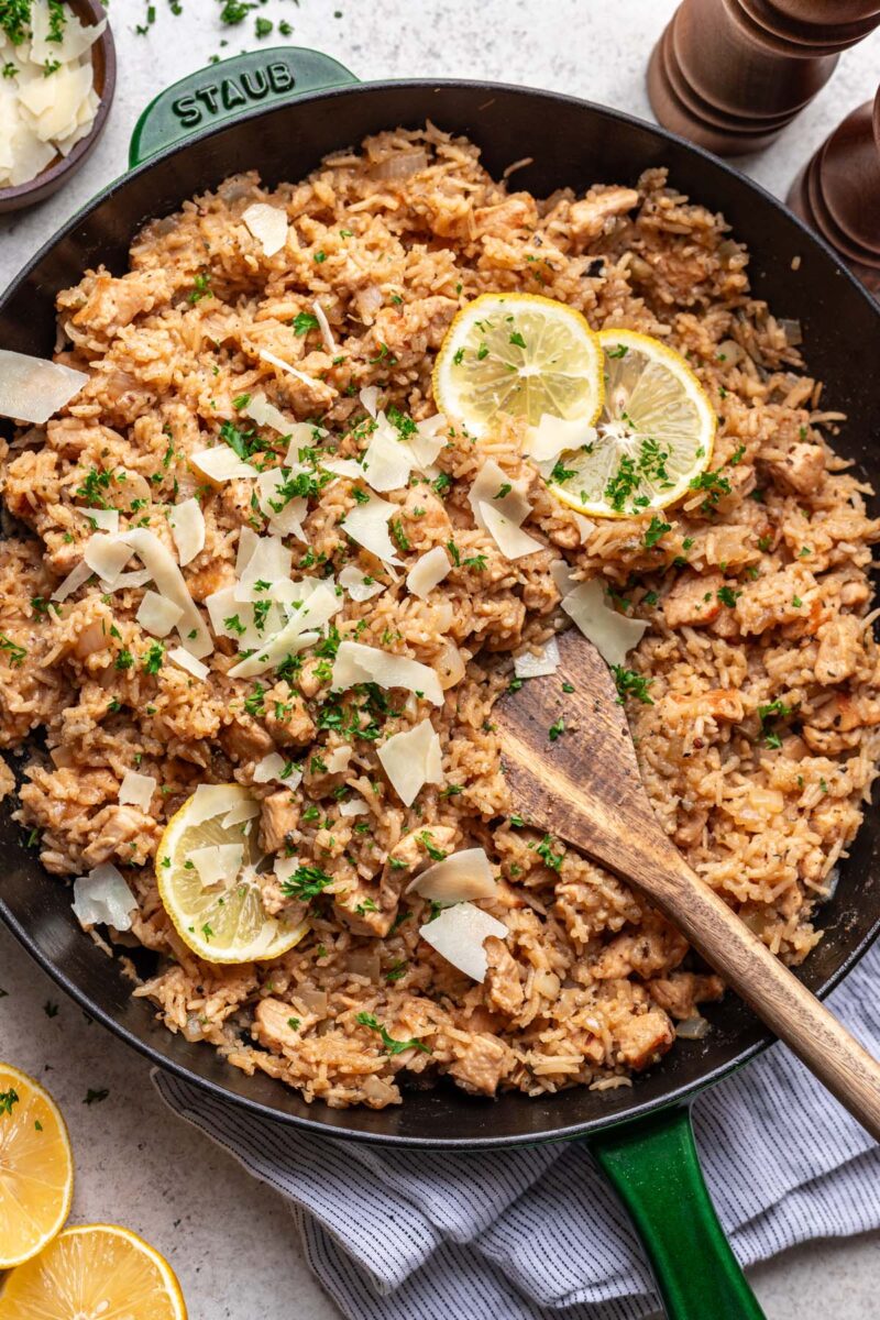 Lemon Parmesan Chicken and Rice in a cast iron skillet garnished with a lemon slice, parmesan and fresh herbs and a wooden spoon in the rice.