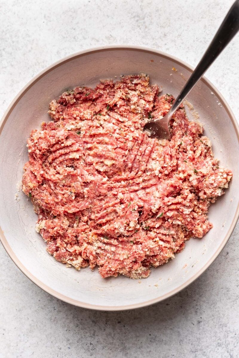 Raw ground meat being mixed together with a fork to make a meatball mixture. 