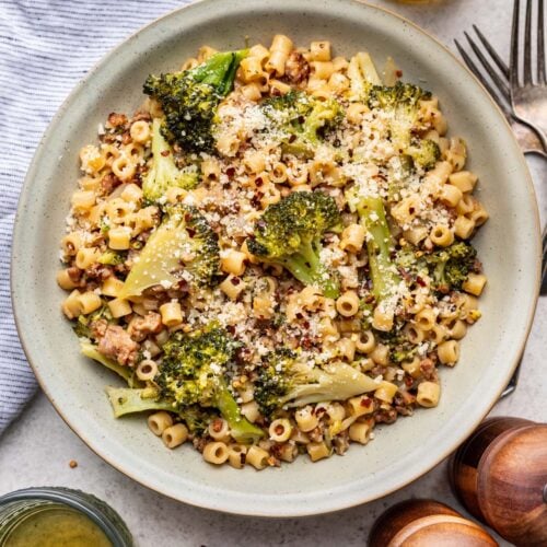 Sausage broccoli pasta in a bowl on a table with salt, pepper, and wine on the side.