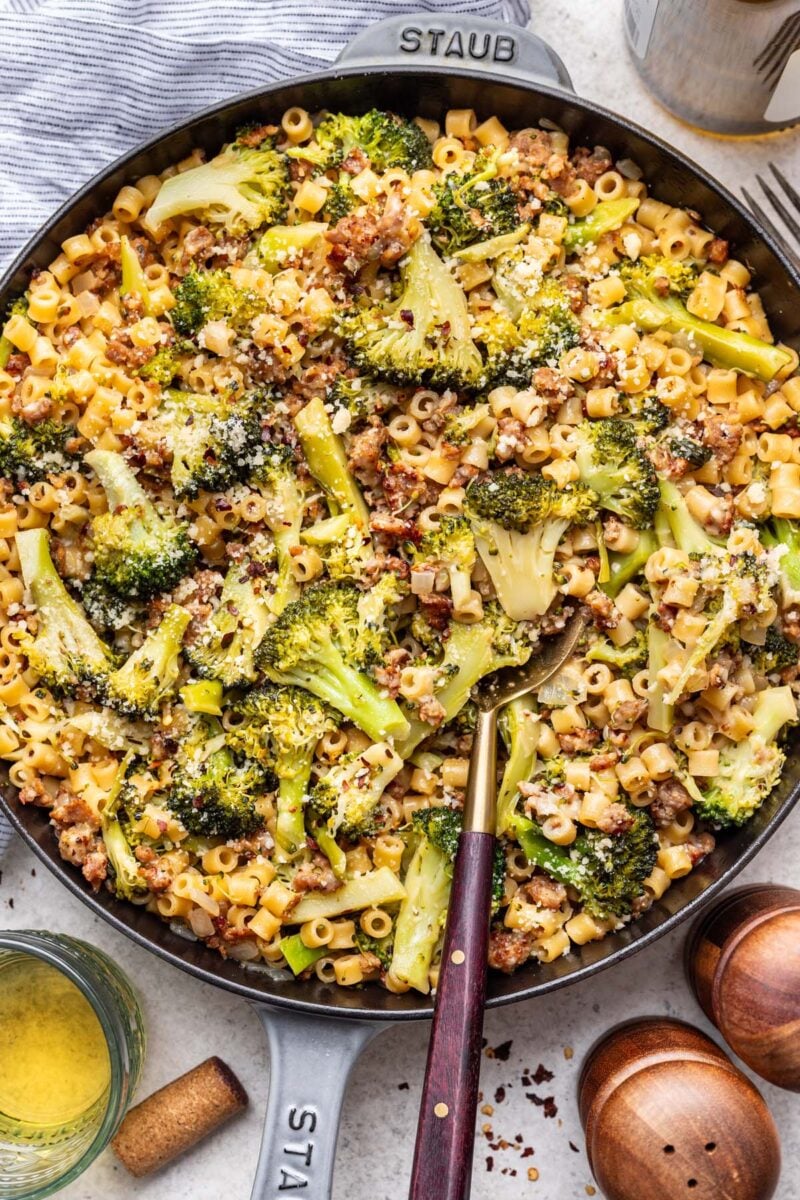 A pan of sausage broccoli pasta with a spoon in it with wine on the side. 