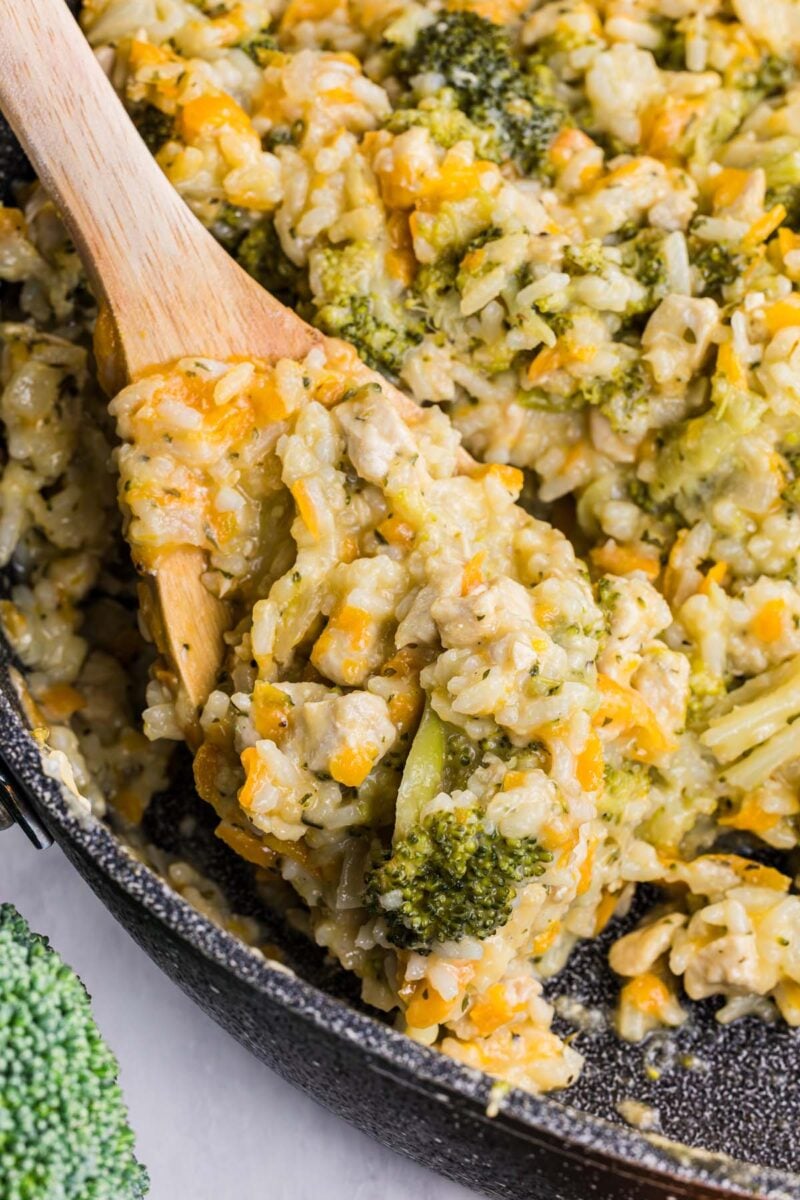 A wooden spoon scooping into broccoli cheddar chicken and rice in a cast iron skillet.