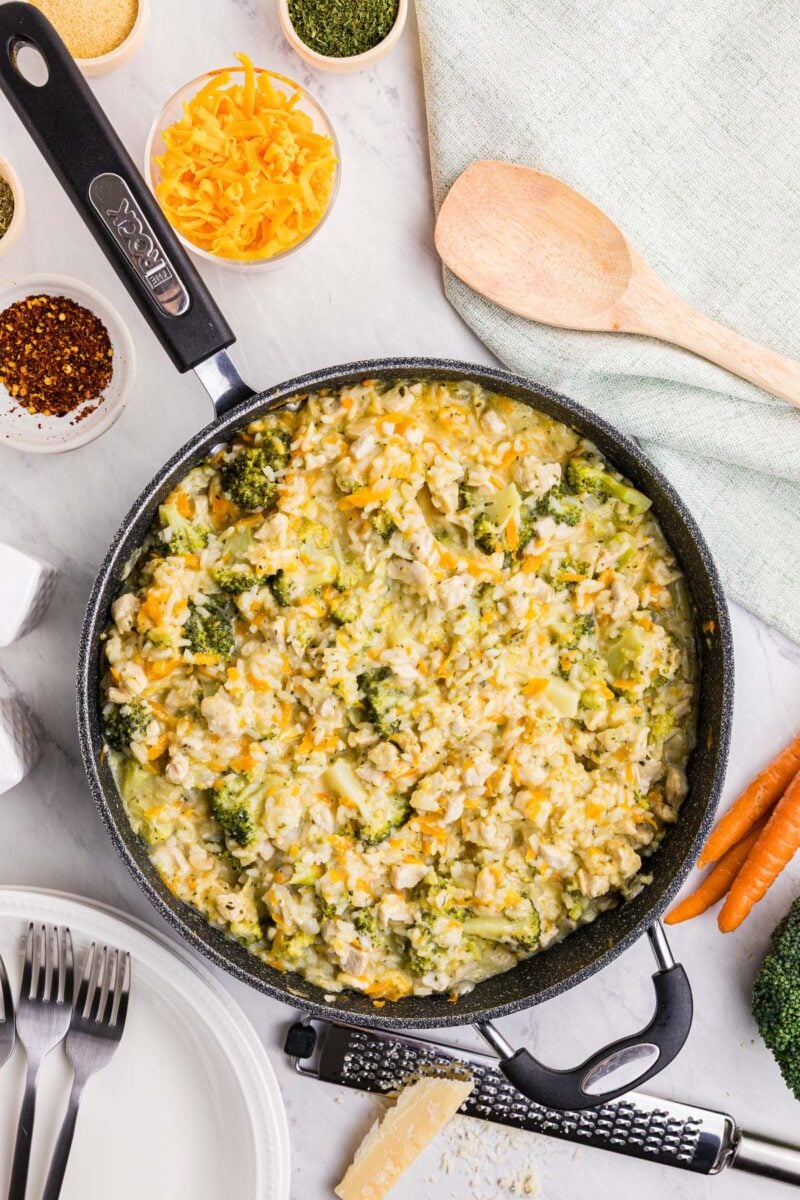 A saute pan with broccoli cheddar chicken and rice and more ingredients on the side. 