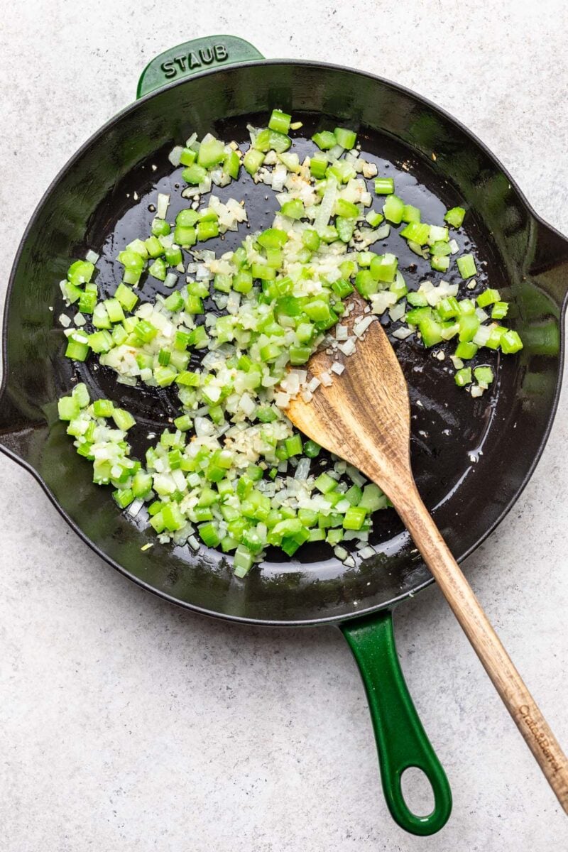 Celery, onions, and garlic sauteeing in a large skillet with a wooden spoon stirring it.