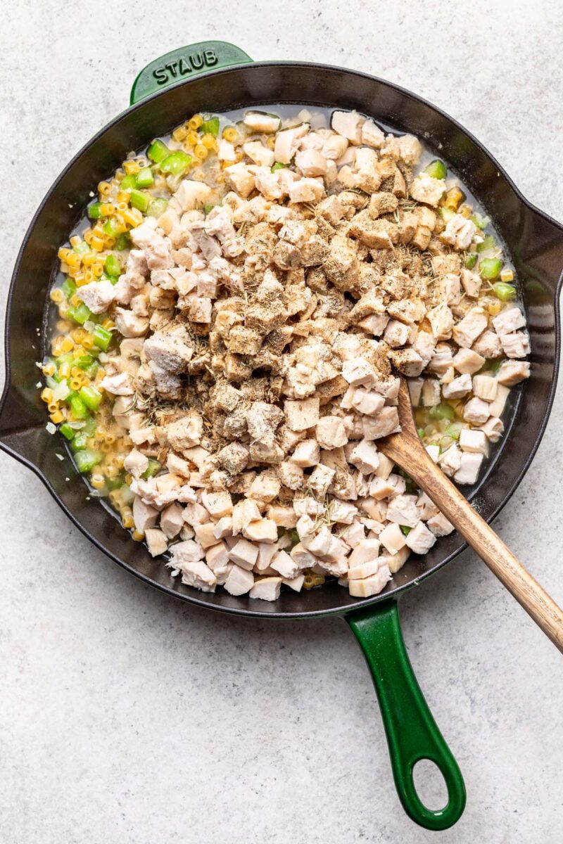 Pasta, celery, onion, garlic, cooked chicken, chicken stock, and spices in a skillet before being mixed together with a wooden spoon in it. 