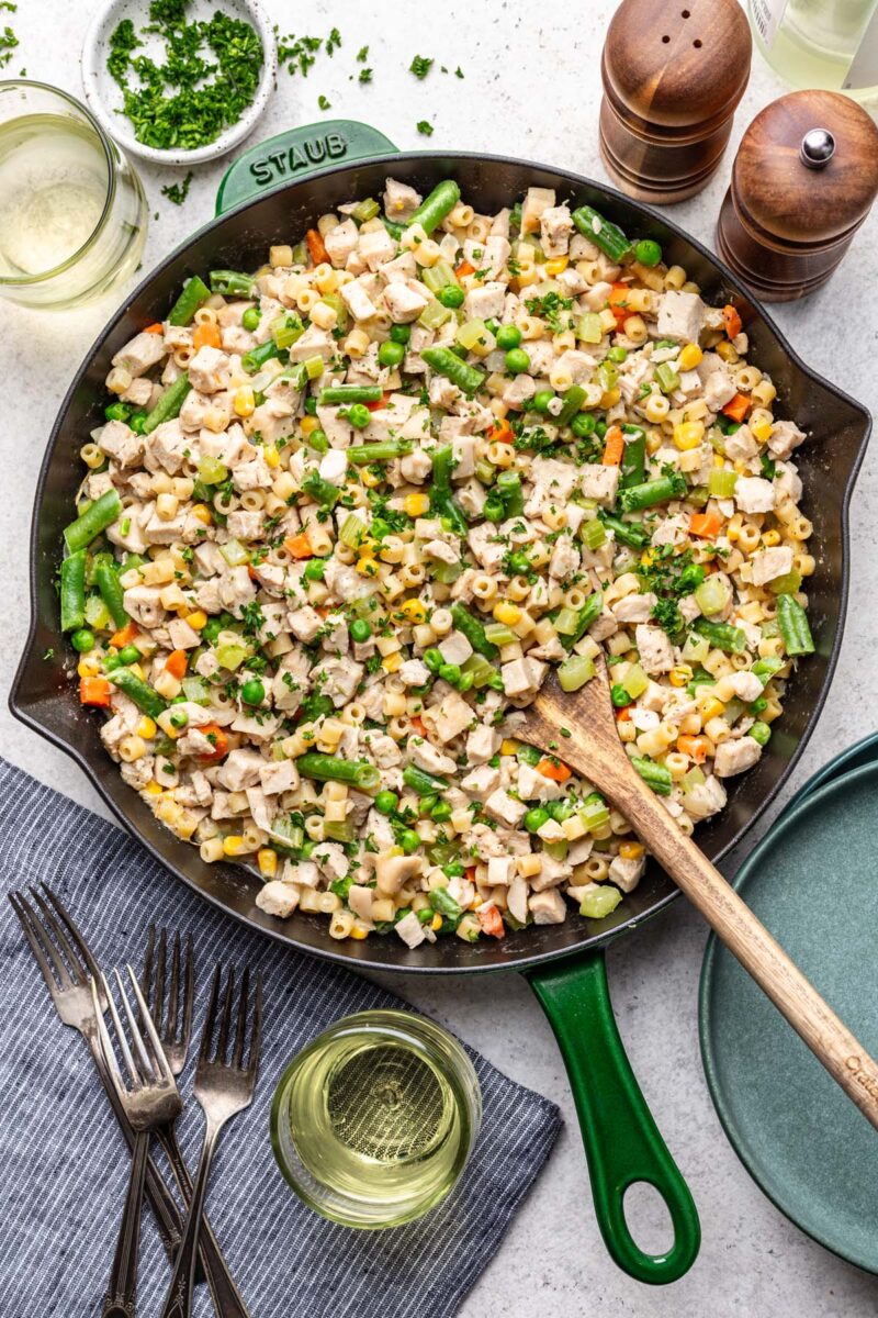 Chicken Pot Pie Pasta in a skillet with a wooden spoon in it with spices and a glass of white wine on the side. 