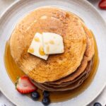 An overhead shot of a stack of oat flour pancakes topped with syrup and 2 pats of butter and a sliced strawberry and blueberries on the side.