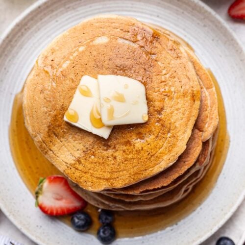 An overhead shot of a stack of oat flour pancakes topped with syrup and 2 pats of butter and a sliced strawberry and blueberries on the side.