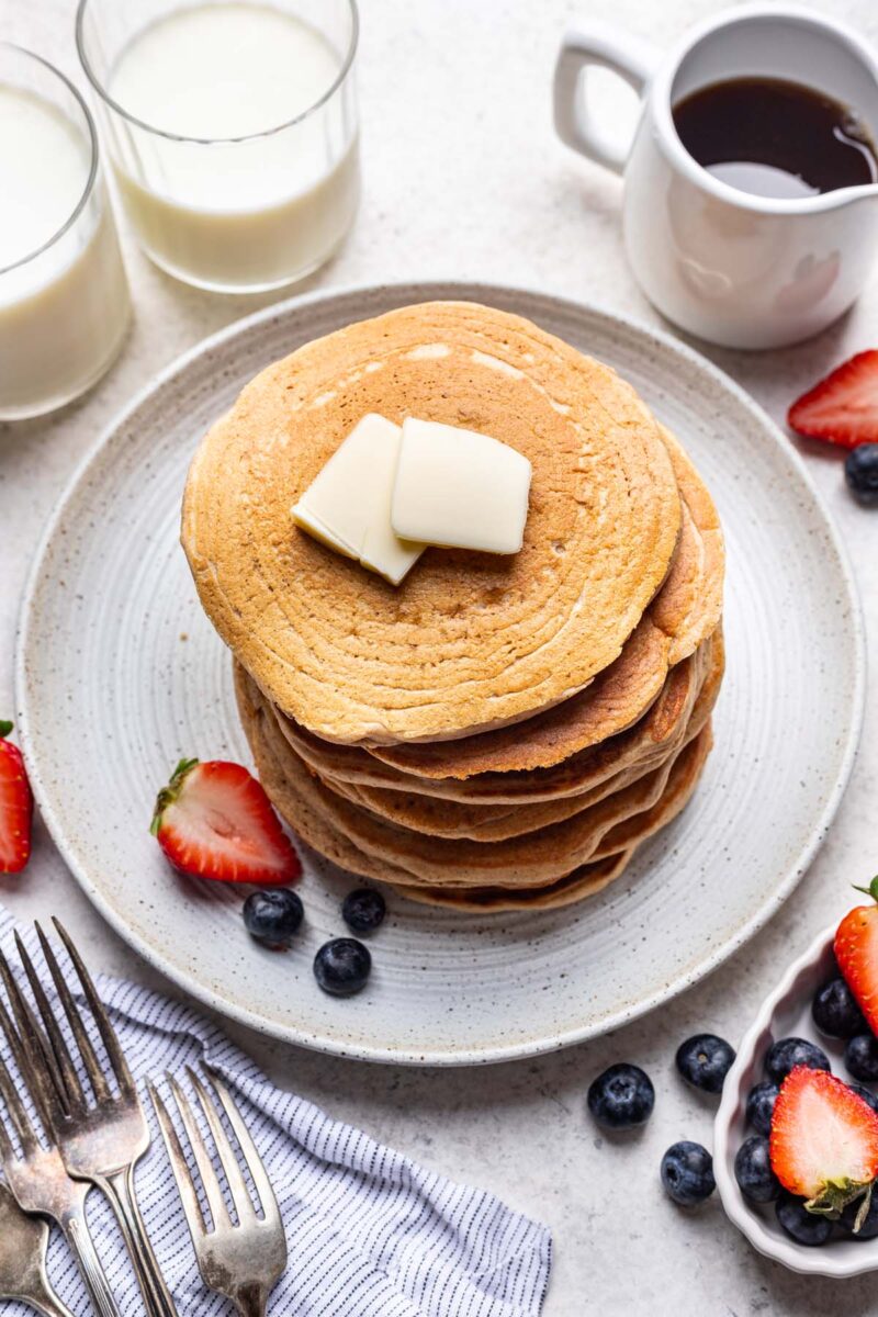 A stack of oat flour pancakes topped with pats of butter and fruit on the side. 